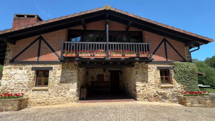 Entrada en piedra del caserío, con balcón de madera y geranios rojos